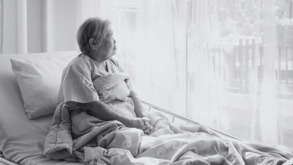 Black and white photo of an older Asian lady sitting up in an hospital bed, looking out the window with a blanket draped over her legs.