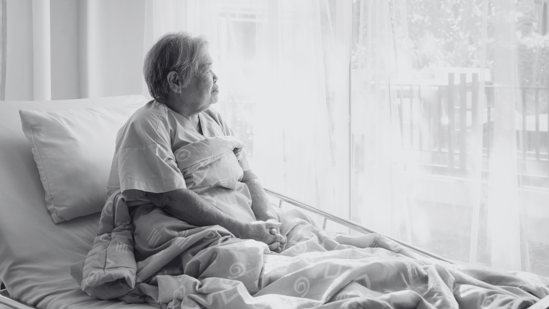 Black and white photo of an older Asian lady sitting up in an hospital bed, looking out the window with a blanket draped over her legs.