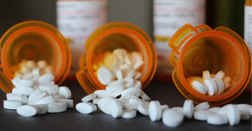 Prescription medication bottles are laying down, lined up, facing the camera. Multiple small, white pills spill out of the containers