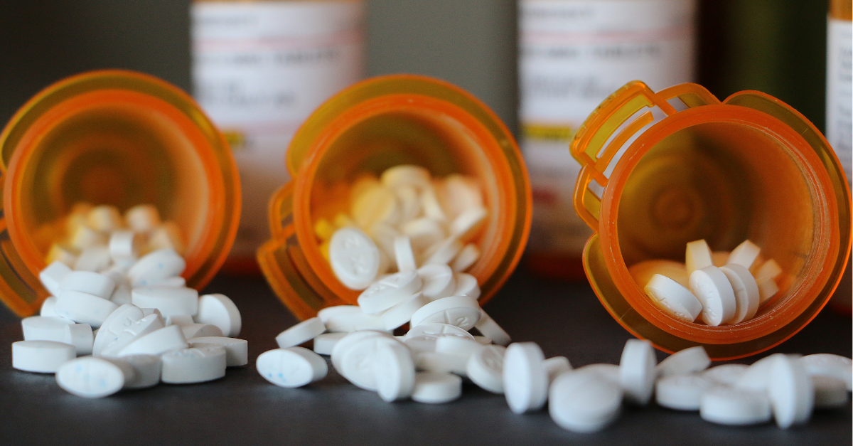 Prescription medication bottles are laying down, lined up, facing the camera. Multiple small, white pills spill out of the containers